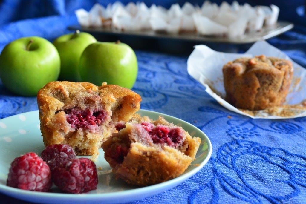 Gluten-free apple and raspberry muffin on a plate with fresh raspberries and green apples in background on a blue tablecloth