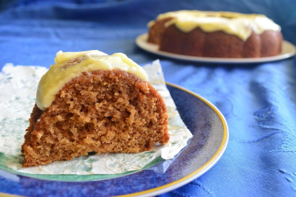 Slice of gluten-free cinnamon teacake with whole cake in background on a blue tablecloth